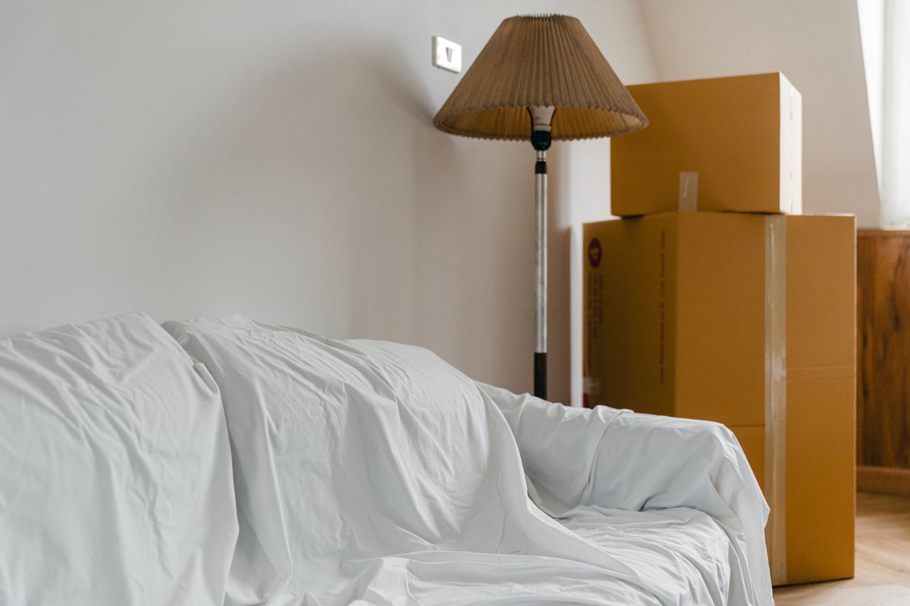 A light, roomy interior of a new contemporary home, with a sheet-covered sofa, a floor lamp, and packed cardboard boxes scattered around.